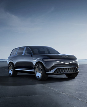 The Genesis Neolun Concept vehicle is parked diagonally in a wide-open field, with its front and right side visible. The top row of two front lights extends horizontally, while the bottom row forms a gentle V-shape at the center of the vehicle’s front.